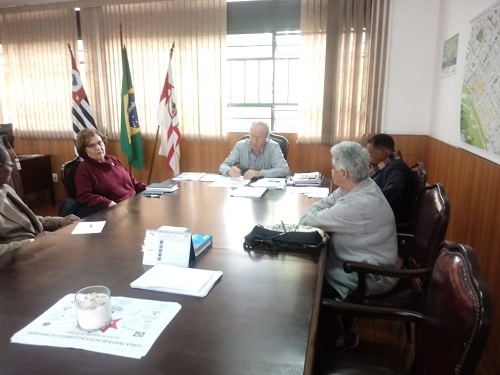 Foto de reunião na sala do subprefeito, com cinco pessoas presentes. Na ponta da mesa encontra-se o subprefeito, ao seu lado esquerdo duas pessoas e ao seu lado direito outras duas pessoas. A mesa de reunião é de madeira, possuí papéis espalhados encima. À esquerda, atrás do subprefeito, há três bandeiras uma do Município de São Paulo, uma do Brasil e do Estado de São Paulo.