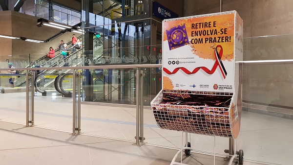 Imagem mostra um dispensador cheio de preservativos masculinos colocado na entrada de um terminal de ônibus, ao lado da escada rolante, próximo ao elevador.