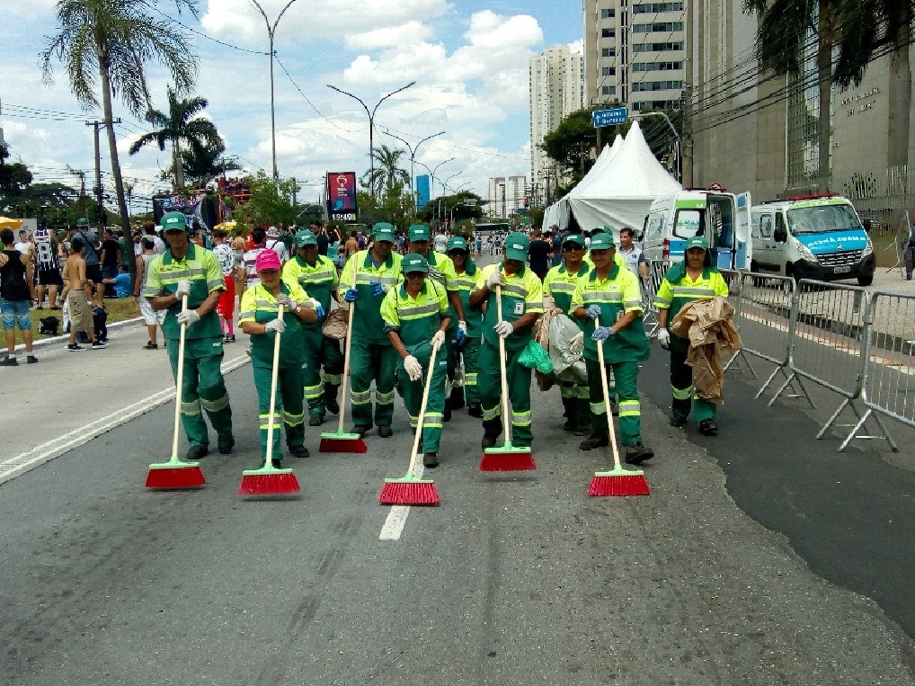 Foto colorida de uma rua onde funcionários de limpeza da Prefeitura, vestidos de verde e segurando uma vassoura, fazem a varrição e limpeza desta rua
