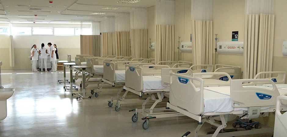 Foto colorida de um leito de hospital com camas hospitalares junto com enfermeiras ao fundo da imagem