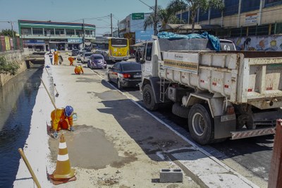 Funcionáros com uniforme laranja tapando buraco no passeio, ao lado direito seis carros estacionados