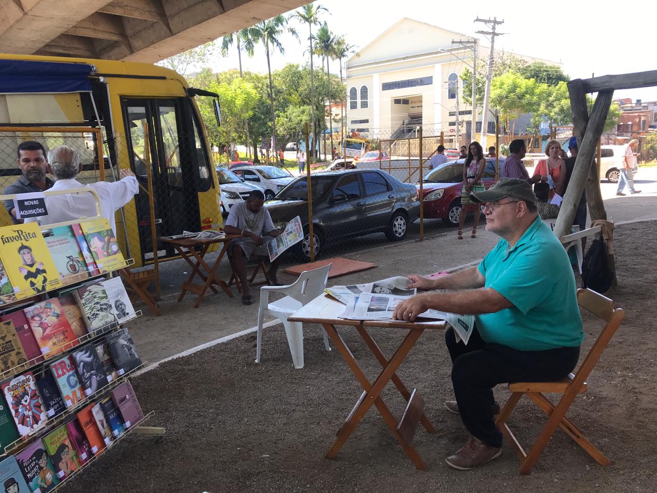 Duas pessoas sentadas lendo jornais e uma estante de livros a esquerda