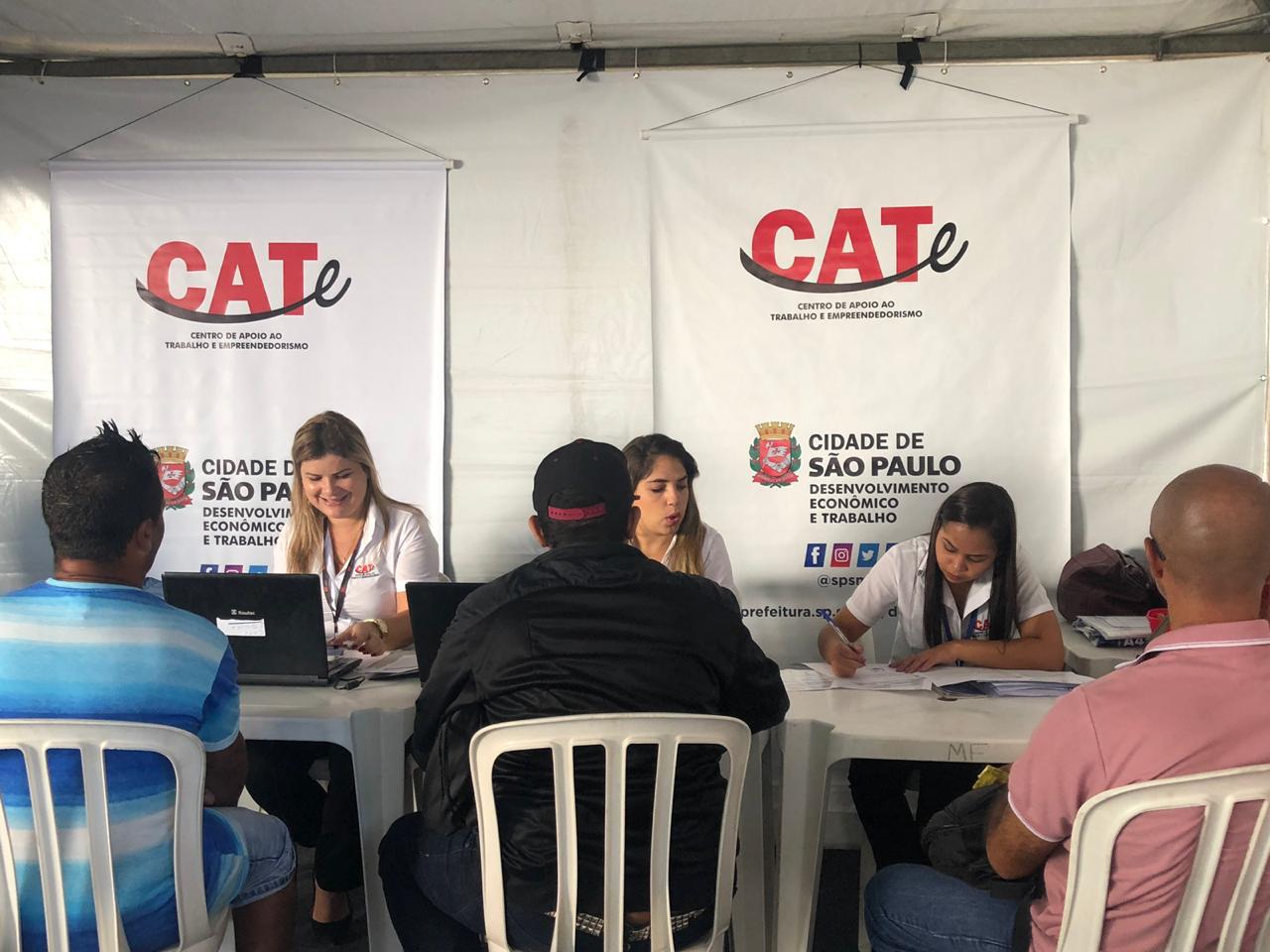 Três atendentes sentadas atendendo três homens. E dois banners do Centro de Apoio ao Trabalhador