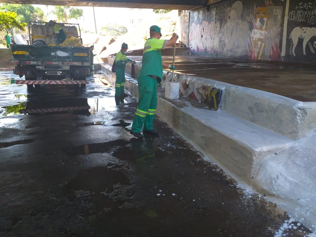 Caminhão com sacos de lixo e três homens uniformizados de verde realizando a pintura, serviço antipichação