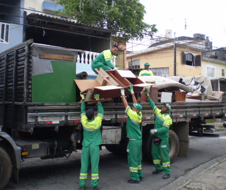 A imagem mostra a entrada de um local que parece ser um ponto de coleta de lixo ou reciclagem, identificado por uma placa com a palavra "ECOPONTO" em letras grandes e vermelhas. A placa também contém informações em português sobre os tipos de materiais que podem ser descartados no local e outras instruções.  A entrada é delimitada por um portão de metal verde, aberto. À esquerda, há uma parede de tijolos pintada de verde com duas faixas horizontais amarelas. A placa do "ECOPONTO" está fixada nesta parede.  À direita da entrada, há uma pequena construção verde com uma porta e uma janela. Mais ao fundo, à direita, observa-se uma área com vegetação em uma encosta. Na entrada, há um carro branco estacionado e dois contêineres amarelos.  O céu está nublado, indicando que a foto foi tirada durante o dia, mas com pouca luz solar direta.