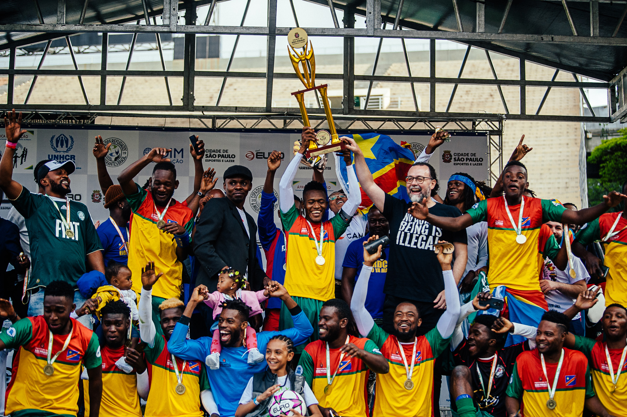 Foto colorida mostra os vencedores da Copa dos Refugiados levantando a taça de campeão