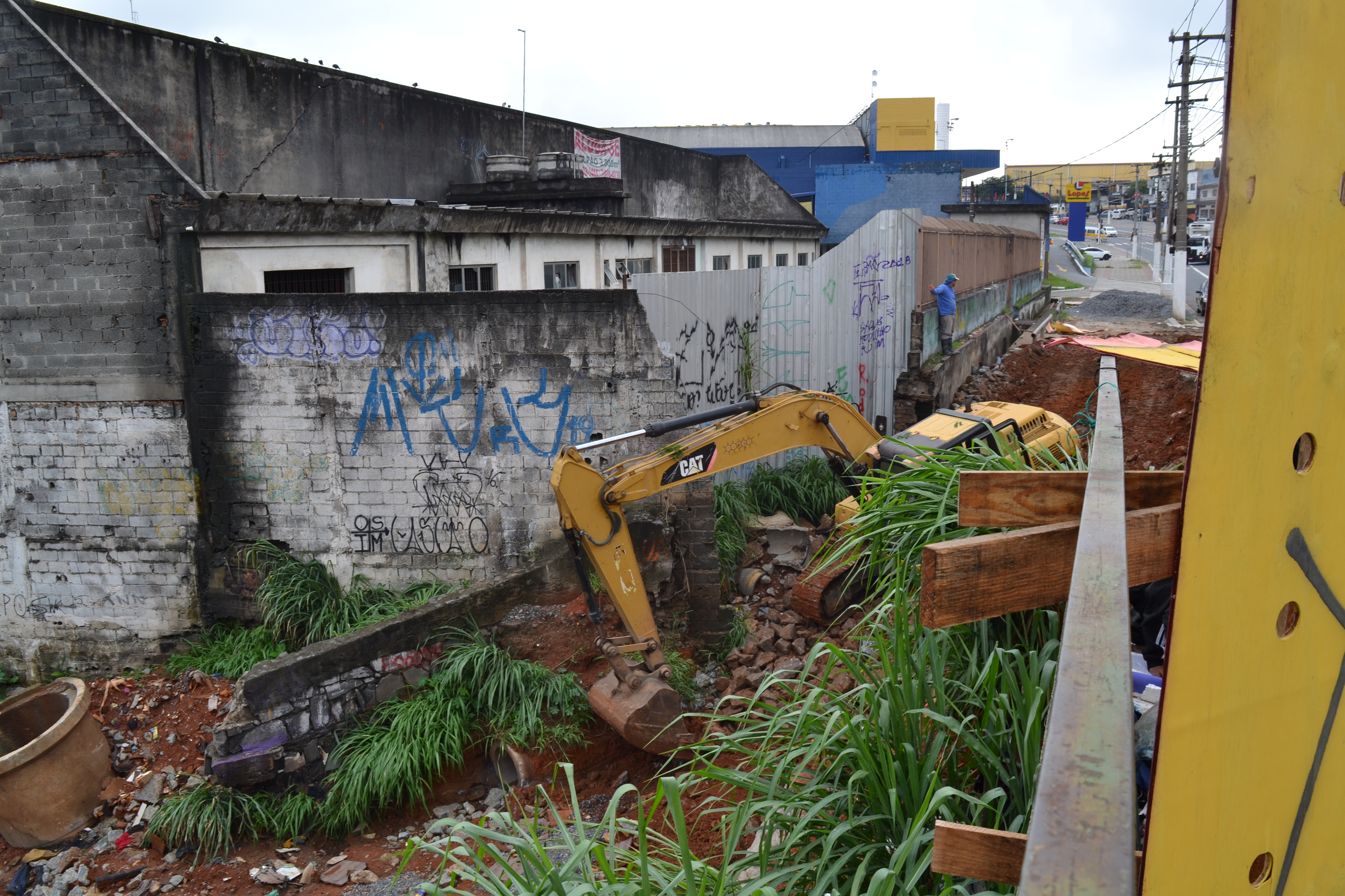 A imagem apresenta um trator limpando a margem do Córrego Itaim, ao lado da ponte na Avenida Marechal Tito. O local contém grande quantidade de entulhos e mato. 