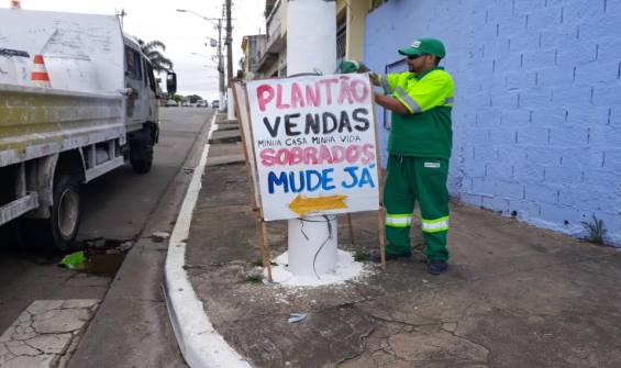 O caminhão de serviços aguarda pelo funcionário que está usando botas pretas, uniforme e boné verde e retirando a placa que está amarrada ao poste ocupando parte da calçada e atrapalhando a passagem do pedestre