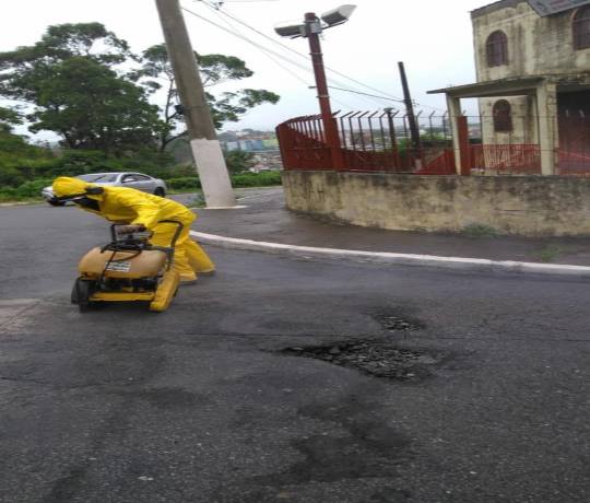 funcionário da SOMA com capa de chuva e capuz amarela, aparelho auricular e máquina tapando buraco 