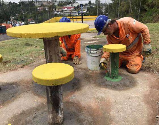 equipe uniformizada faz pintura do mobiliário urbano . Mesa e banquinhoa em amarelo e pezinhos na cor verde. Ao fundo a quadra já pintada em uniformidade com a cor amarela.