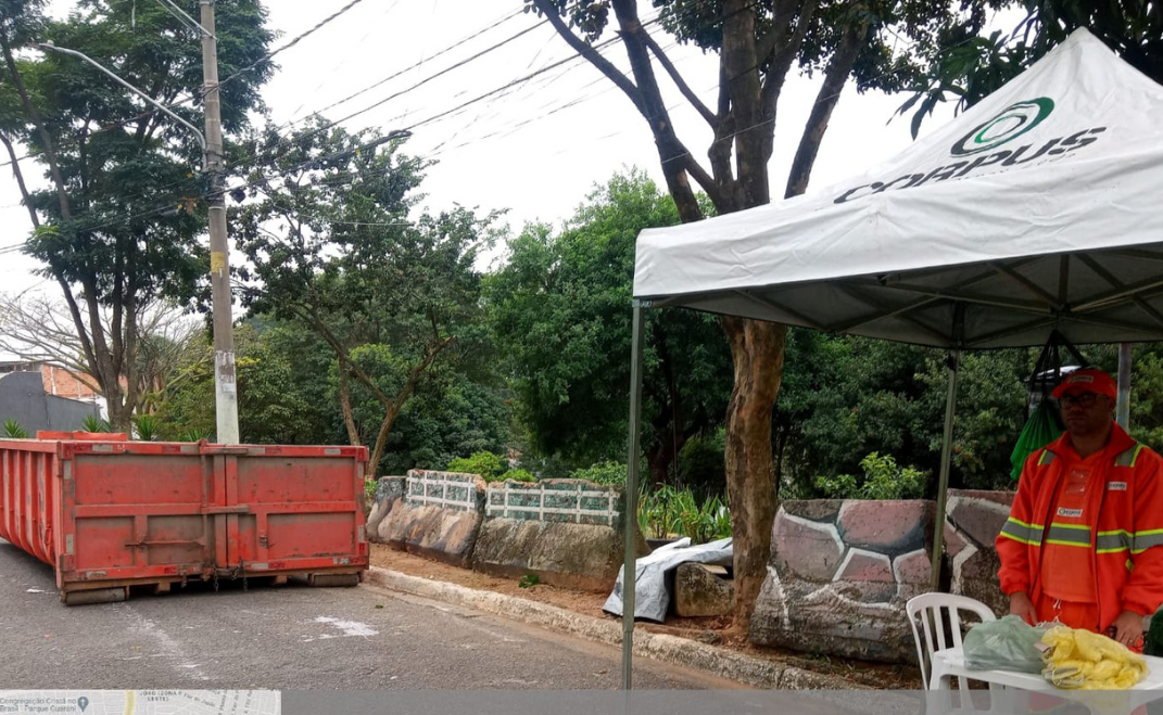 A imagem mostra uma tenda branca montada em uma área externa, possivelmente em uma rua ou calçada. A tenda tem o logotipo "CYCLUS" impresso no topo, em letras verdes e um símbolo circular também em verde.  Sob a tenda, há um homem usando um uniforme laranja com detalhes em amarelo e um capacete laranja. Ele está sorrindo e olhando para a câmera. Ao lado dele, há uma cadeira branca e uma sacola amarela no chão.  À esquerda da tenda, há um grande contêiner de lixo vermelho sobre um palete de madeira. Ele está posicionado na rua, próximo à calçada.  Ao fundo, há vegetação, árvores e um muro de pedras. O ambiente parece ser um dia nublado.  A cena sugere um evento ou ação relacionada à coleta de lixo ou reciclagem