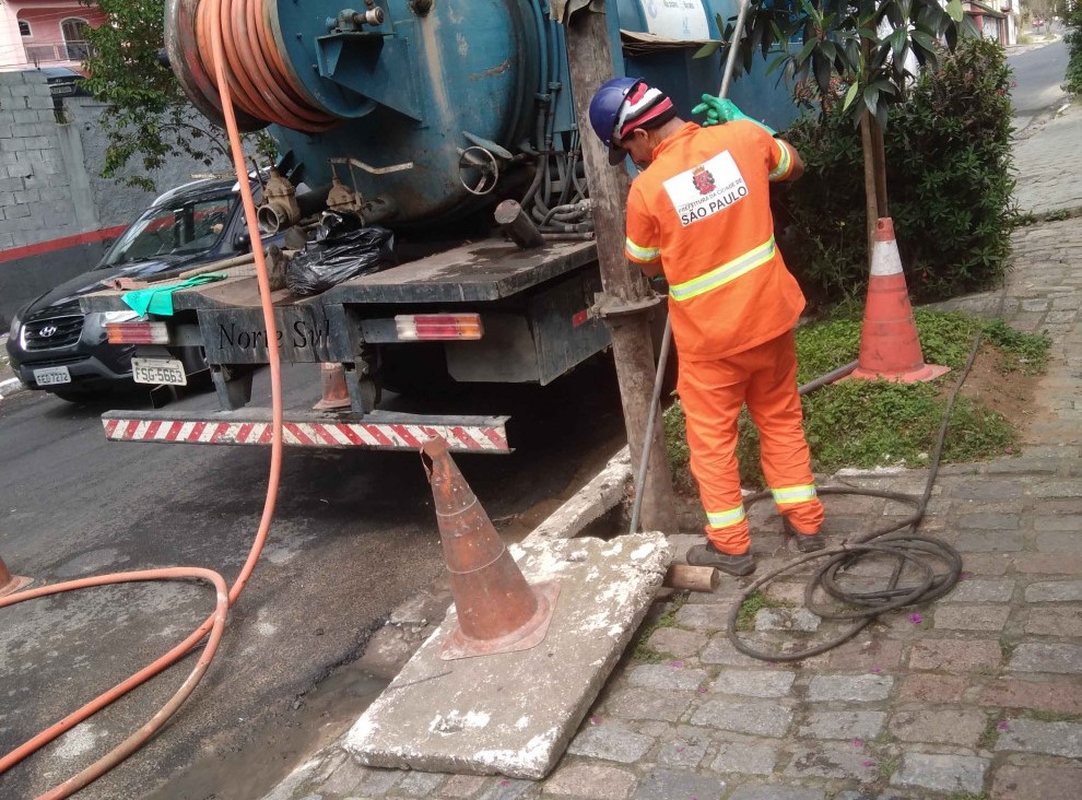 A imagem mostra um caminhão de cor azul claro, com um grande tanque na carroceria. O caminhão está estacionado em uma rua de paralelepípedos.  Um homem, usando um uniforme laranja com detalhes em amarelo e um capacete de segurança, está trabalhando próximo a uma árvore e a uma boca de lobo na calçada. Ele está de costas para a câmera e parece estar manipulando uma mangueira laranja que sai do caminhão.  Há também um carro preto estacionado parcialmente na rua, ao lado do caminhão.  Dois cones de sinalização laranja estão no chão, indicando a área de trabalho. Um está próximo à boca de lobo e o outro mais distante, na direção do caminhão.  O ambiente parece ser uma rua residencial ou comercial, com árvores e algumas construções ao fundo. O dia parece estar ensolarado.  A cena sugere que o homem está realizando algum tipo de serviço de manutenção ou limpeza, possivelmente relacionado a encanamento ou esgoto.