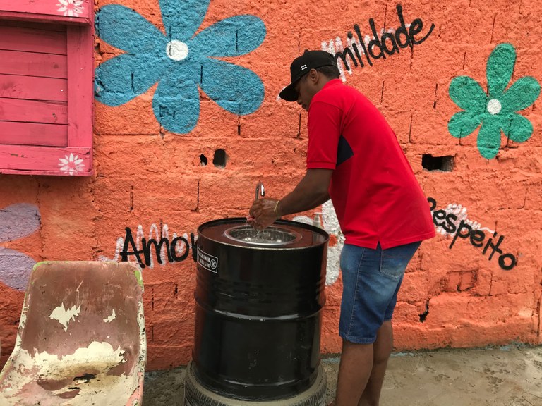 imagem mostra-se a pia com o formato um barril preto e um pneu em sua base e os moradores da regioão utilizando a pia para se higienizar.