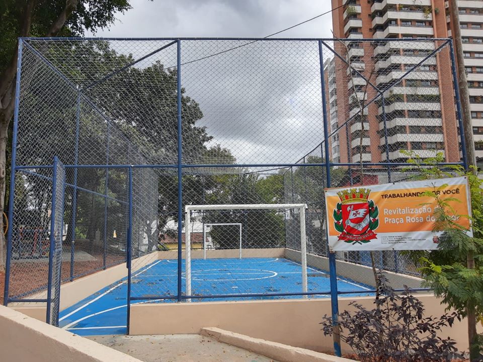 Imagem com obtem a quadra no centro a mesma possui piso na cor azul e grades no seu entorno e em sua frente contém a placa na cor laranja e branca com as informações sobre a obra.