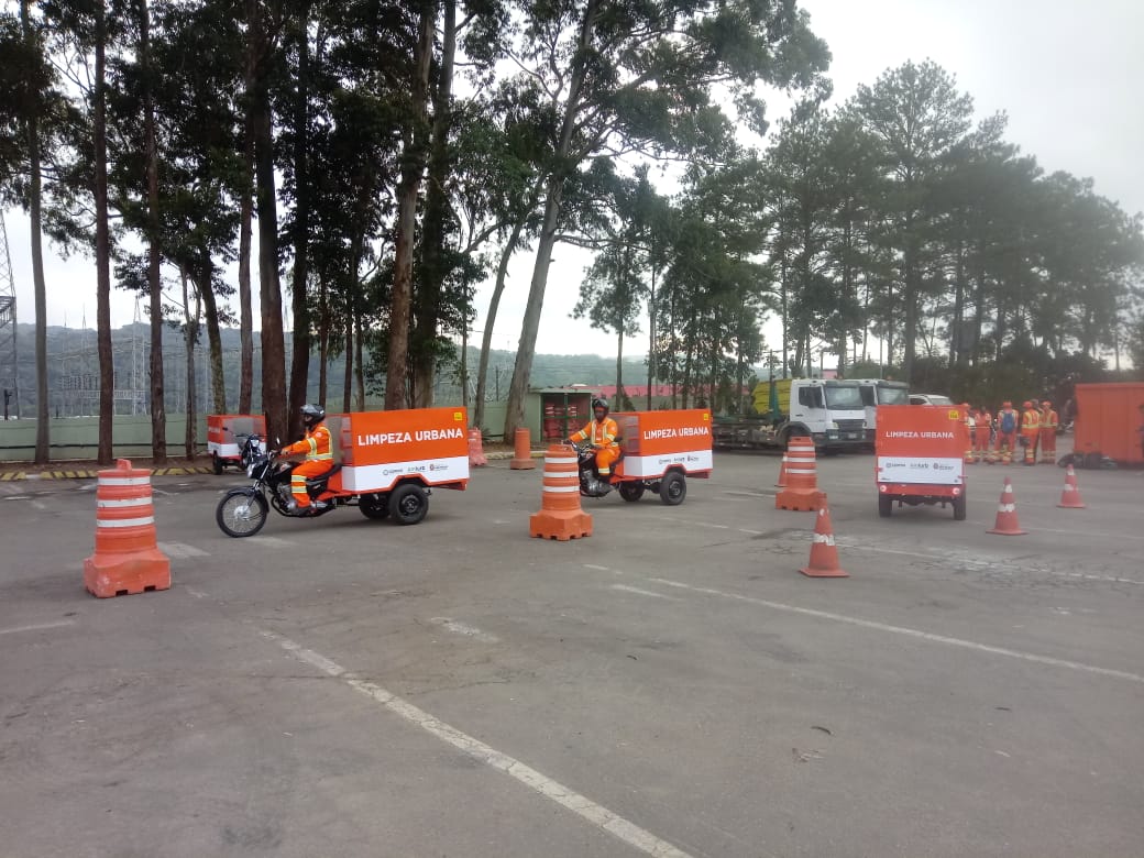 Motociclistas uniformizados fazem manobras entre cones, com triciclos da limpeza, de cor laranja.