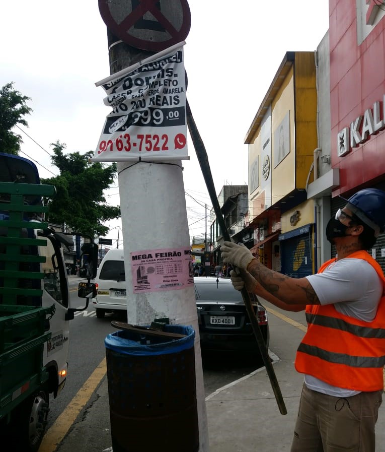 Funcionário da Prefeitura com uma foice na mão, retira placa irregular de um poste