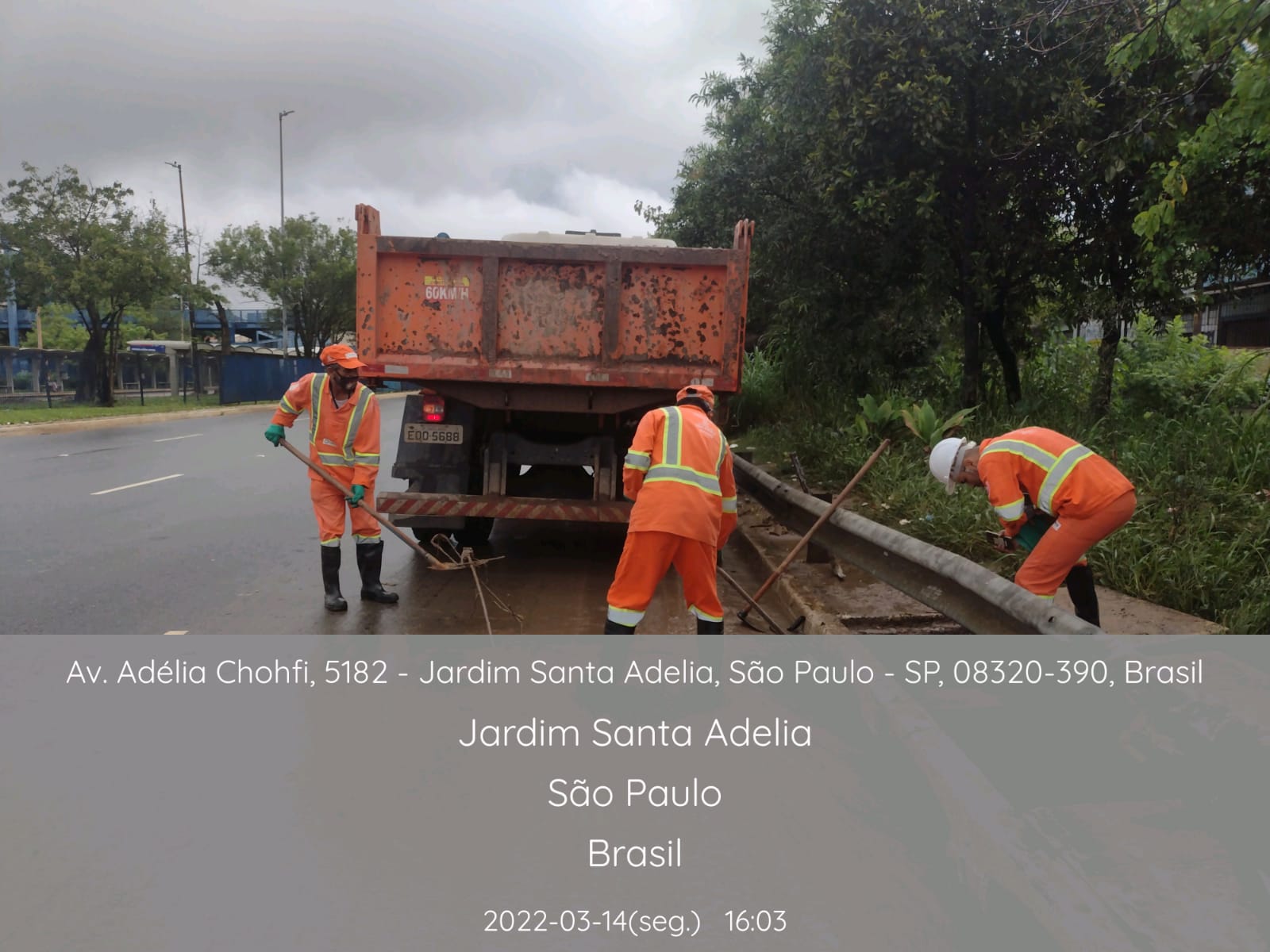 Três funcionários da Prefeitura, com uniforme laranja,. jogam lama dentro de um caminhão basculante, usando pás. 