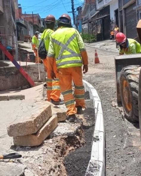 Trabalhadores montam guias e sarjeta em uma rua. Vestem camisão reflexivo, na cor verde limão e capacetes marrom e vermelho. 