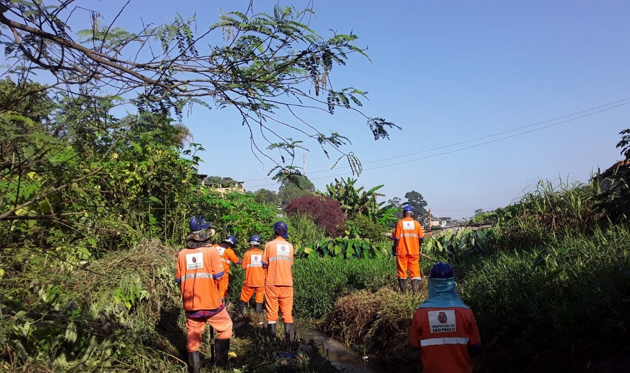 4 Trabalhadores localizados no centro da imagem, eles usam uniformes laranjas e ferramentas para ajuda na limpeza do córrego localizado abaixo deles, em sua volta um grande matagal. 
