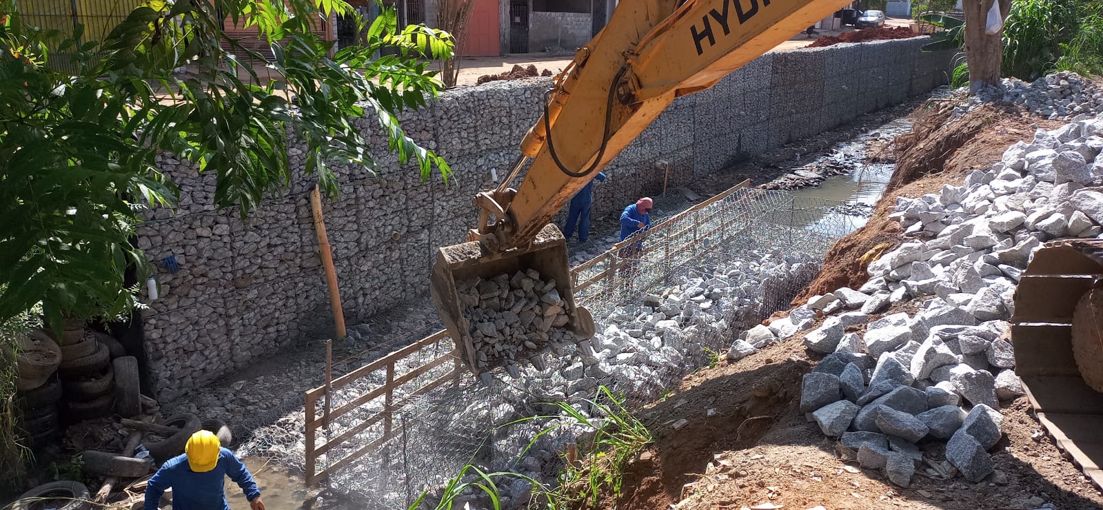Escavadeira move pedras, enquanto trabalhadores terminam a obra.