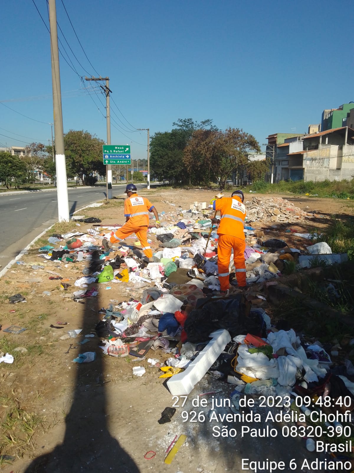 Dois homens com uniforme laranja trabalham na limpeza de um ponto de descarte irregular de lixo e entulho.