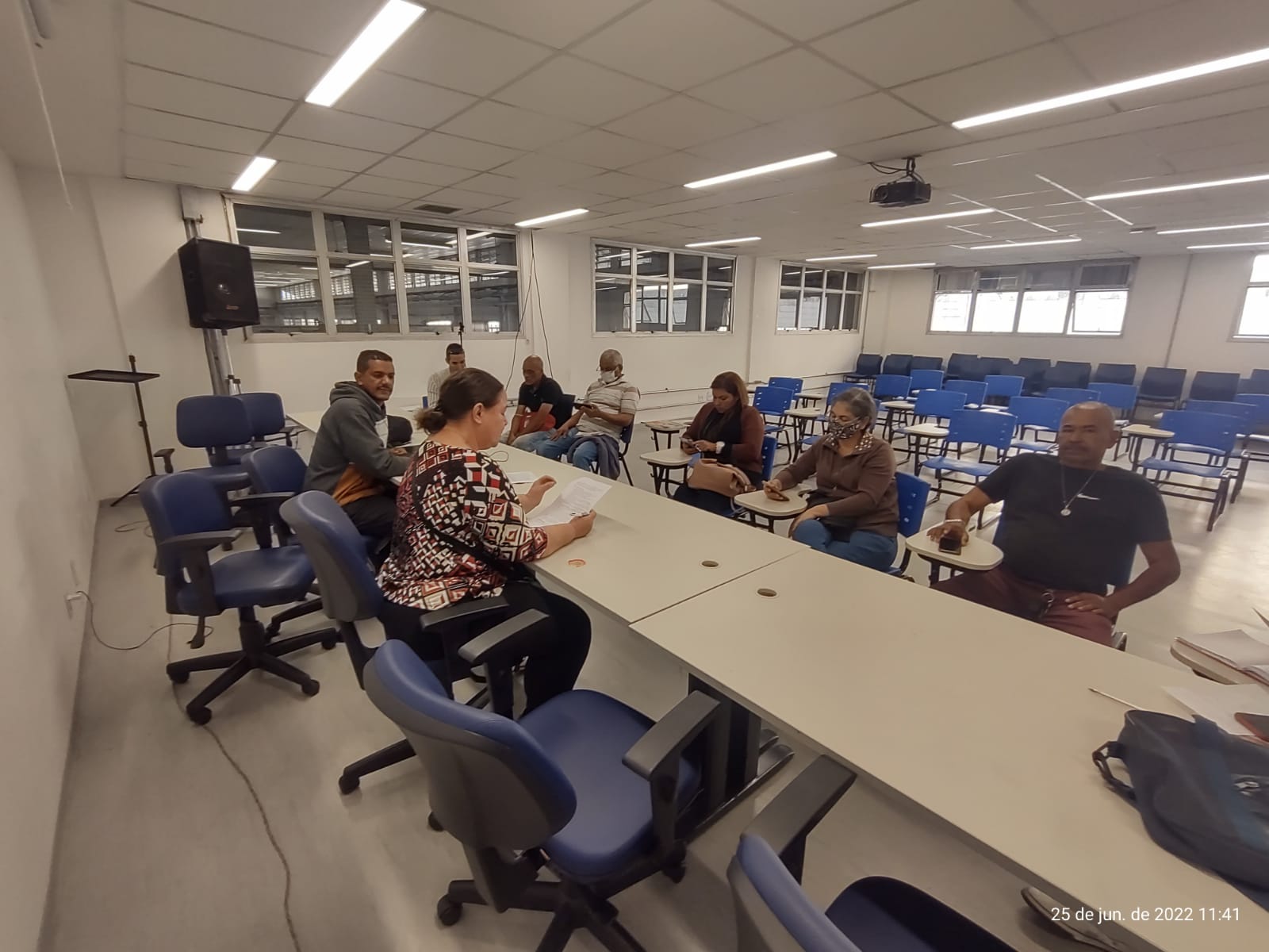 à frente de uma mesa, uma mulher e um homem, conduzem uma reunião, com mais seis pessoas sentadas à frente. 