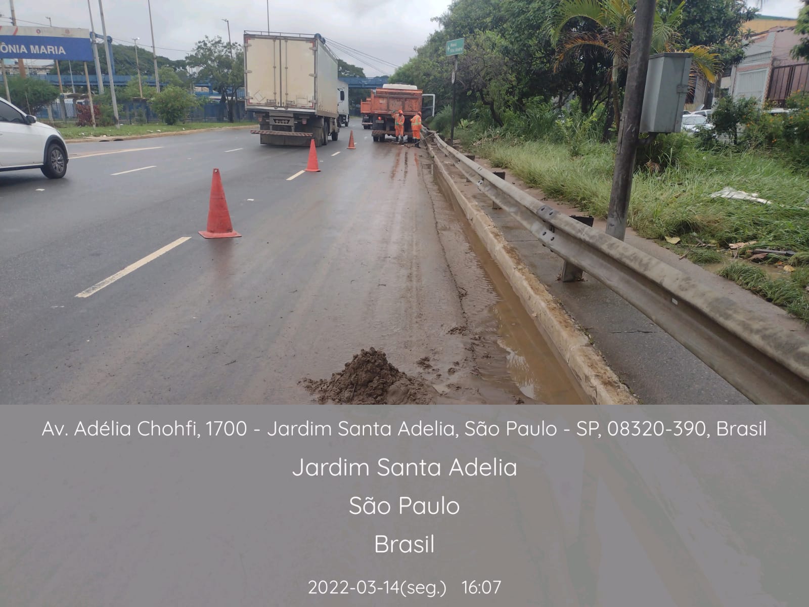 Cones de sinalização estão dispostos em uma faixa de tráfego da Avenida Adélia Chohfi. Ao fundo, trabalhadores limpam a pista, atrás de um caminhão basculante. 