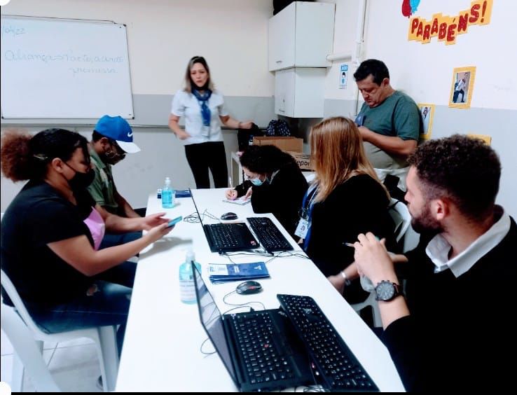 Sala de atendimento, duas pessoas sentadas com computadores em sua frente, e outras do outro lado da mesa sendo atendidas.