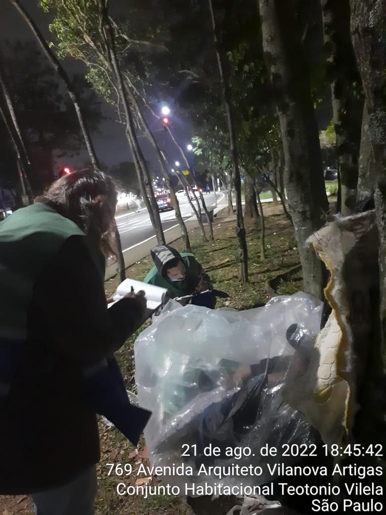 Duas pessoas com jaleco da Assistência Social, estão dialogando com uma pessoa em situação de rua, que está deitada em um gramado, enrolada em pedaços de plástico e cobertor r´pustico