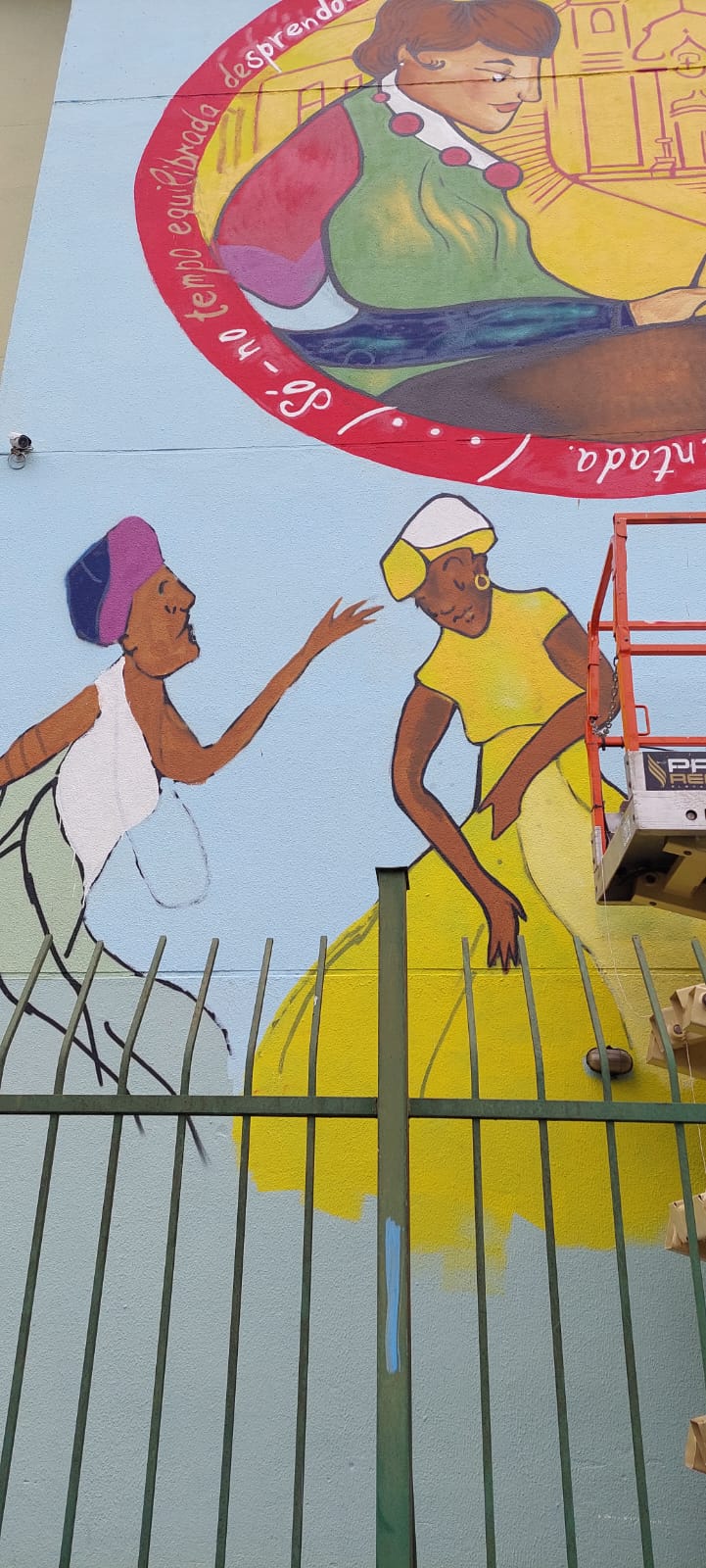 Jovem pintora trabalha em cima de um andaime, finalizando a fachada de um prédio, pintado com representações humanas, realizando trabalhos domésticos. 