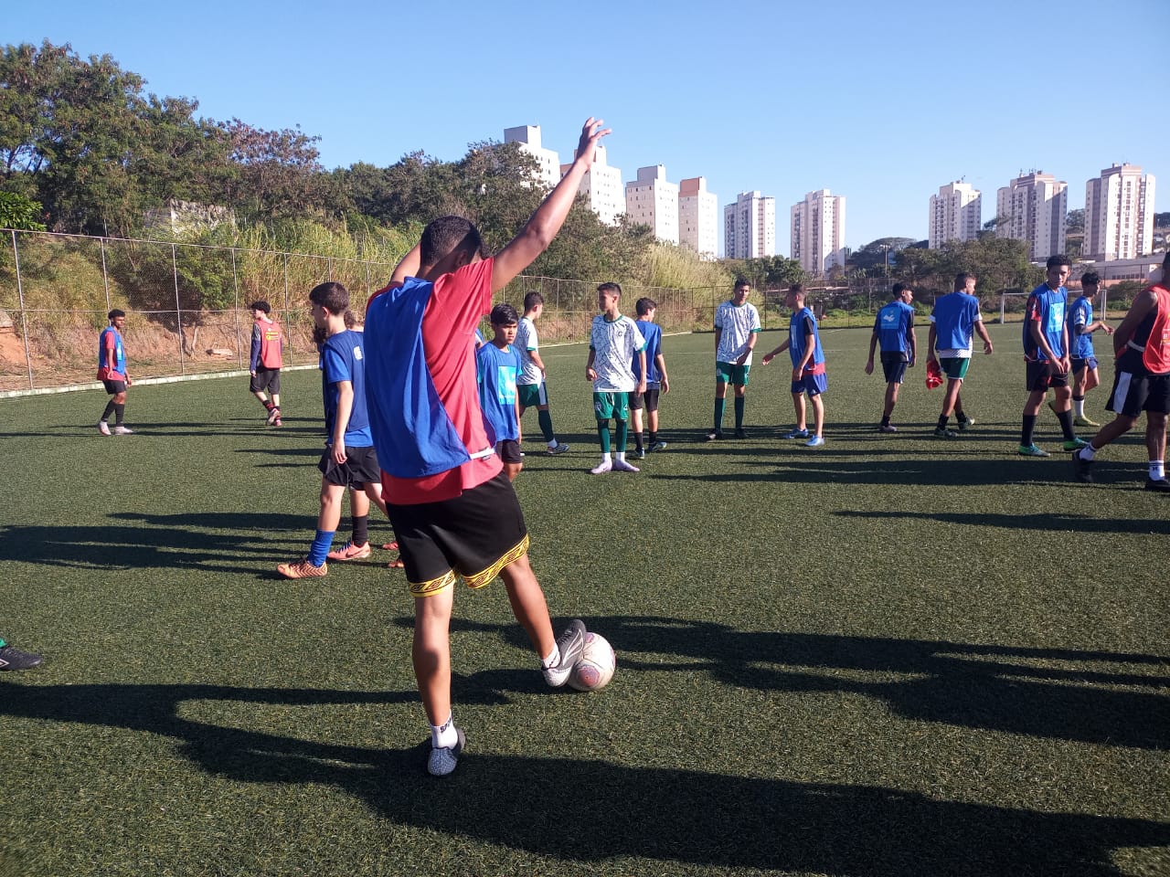 Em um campo de futebol com gramado sintético, um jovem ergue o braço, sinalizando para outros jogadores que vai chutar. 