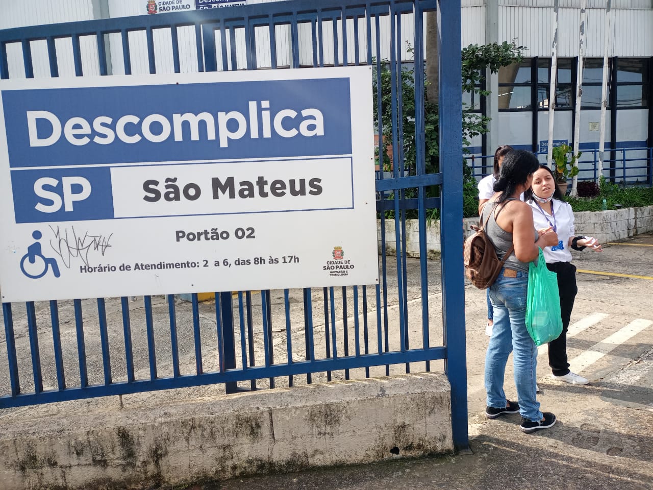 Placa do Descomplica São Mateus afixada no portão com grades de ferro, indicando portão de entrada 02, horário de atendimento, de segunda a sexta, das 8h às 17h. à esquerda, o símbolo de local acessível. 