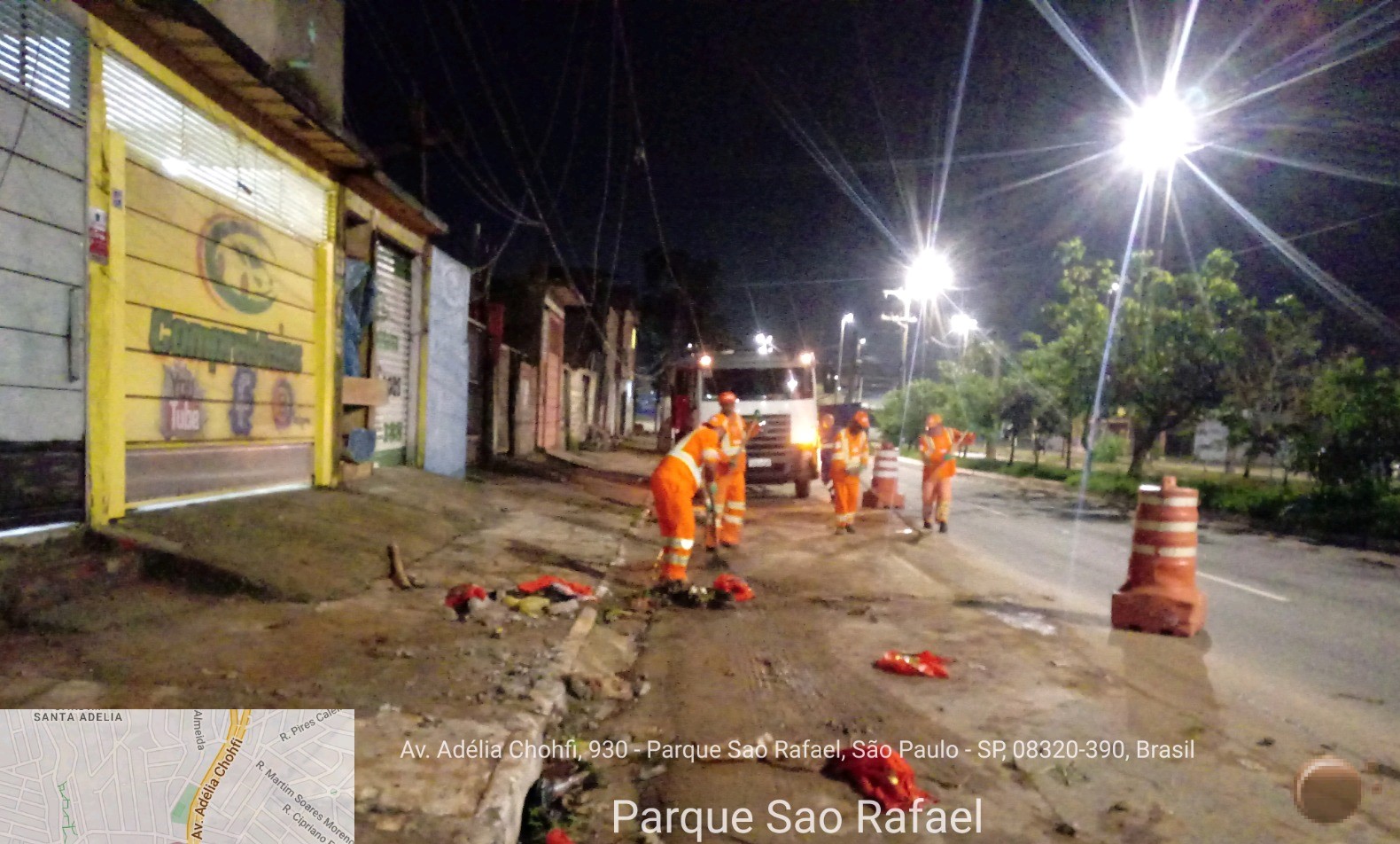 Trabalhadores vestidos com macacão laranja retiram lama da pista em uma avenida iluminada. Cones isolam a faixa que está suja. 