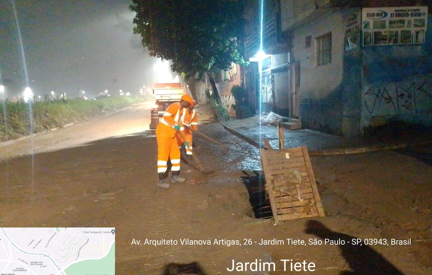 Trabalhadores com uniforme laranja limpam rua com barro, em frente a residências. Foto identificada como Av. Vilanova Artigas, Jardim Tietê.