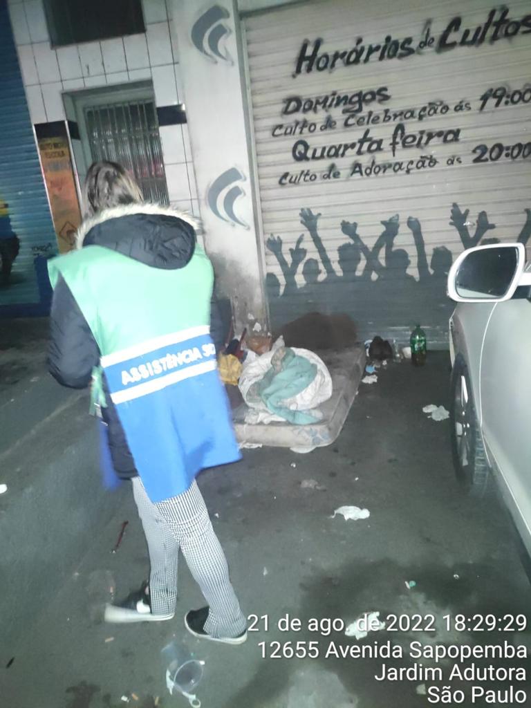 Mulher com jaleco da Assistência Social, está diante de uma pessoa em situação de rua deitada no chão, enrolado em um cobertor rústico. 