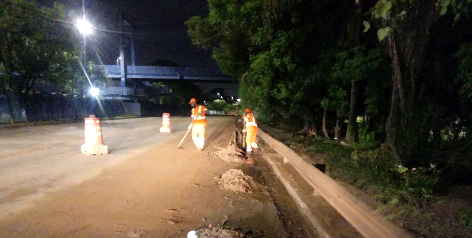 Dois trabalhadores, com macacão laranja limpam a lama de pista, com enxadas. Cones isolam a área afetada. 