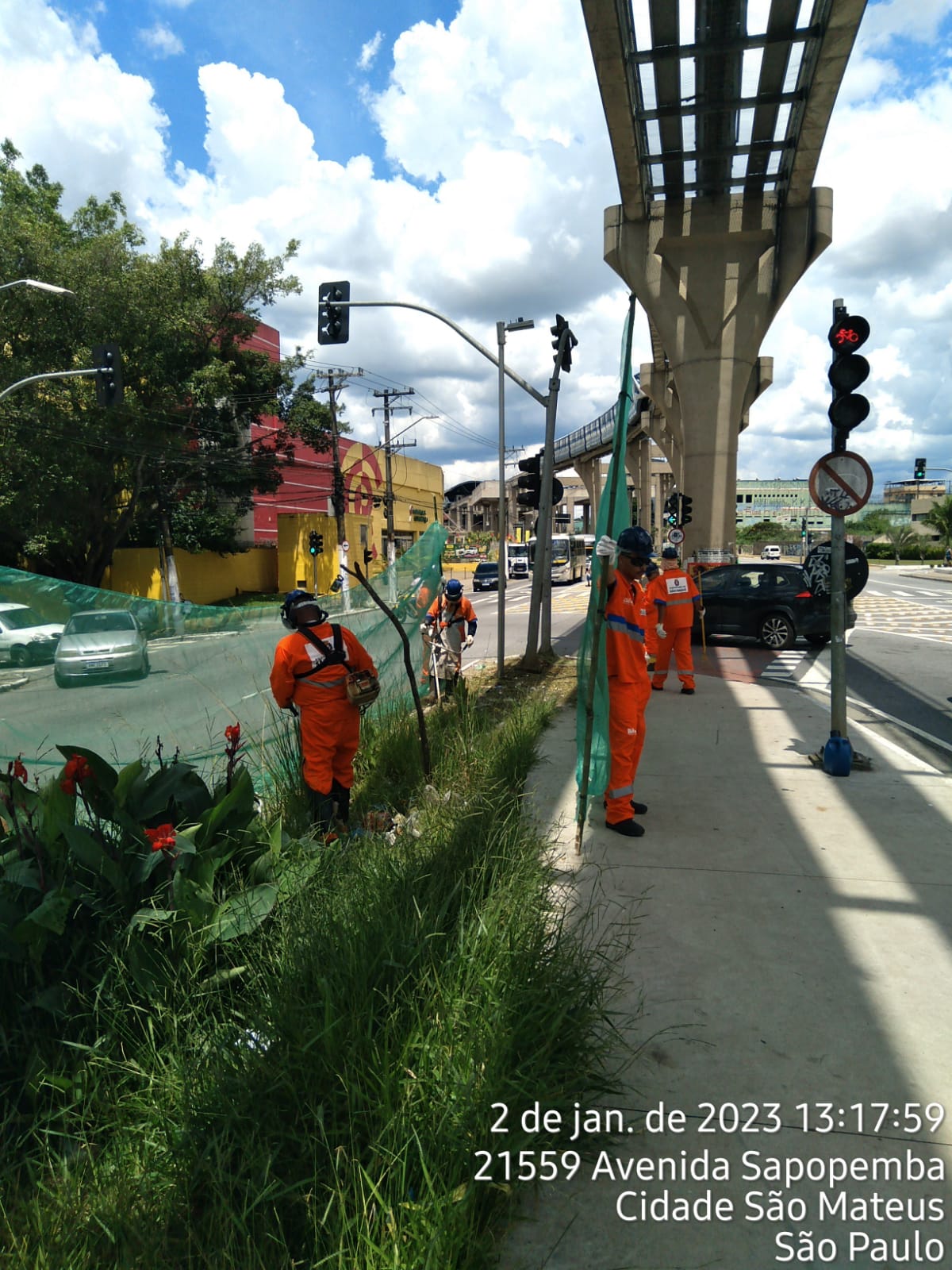 Funcionários da Prefeitura, com uniforme laranja, fazem o corte de grama na Avenida Sapopemba, altura do 21.559, embaixo da linha do Monotrilho.