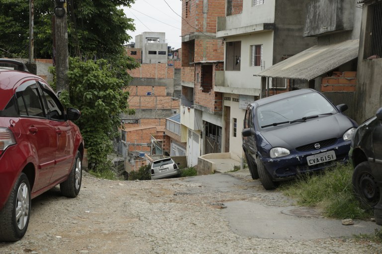 Em uma viela estreita, dois carros estão parados nas laterais e um terceiro desce uma ladeira, em que só é possível enxergar o teto do veículo. Embaixo, várias casas sem reboco externo. 