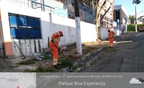 Dois trabalhadores com uniforme laranja, fazem capina de mato sobre a calçada em uma rua. 