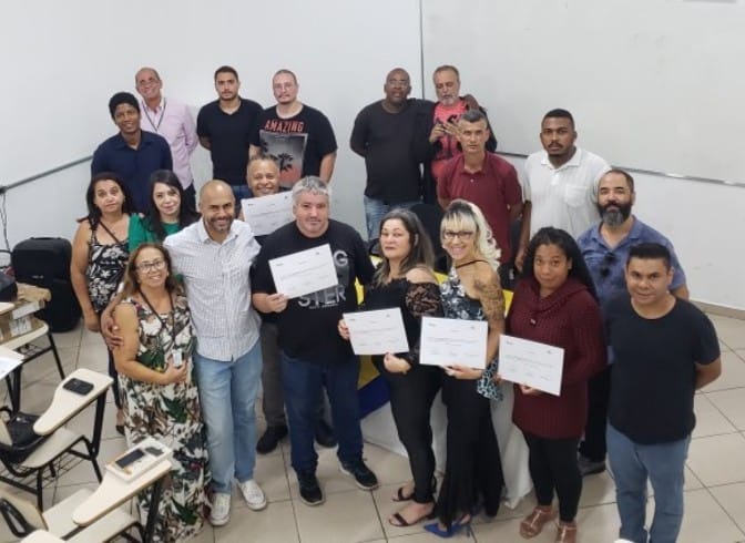 A foto mostra um grupo de aproximadamente dezoito pessoas adultas, de diversas aparências, reunidas em um ambiente interno, posando para a câmera com sorrisos e expressões de satisfação. A maioria segura um certificado branco em suas mãos.  As pessoas estão dispostas em duas fileiras. Na frente, há um grupo mais numeroso, com homens e mulheres lado a lado, segurando seus certificados voltados para a câmera. Na fileira de trás, há alguns homens em pé, também sorrindo.  Os certificados são retangulares e brancos, com alguma inscrição preta visível, sugerindo que acabaram de receber algum tipo de reconhecimento ou conclusão de curso.  As vestimentas são variadas, incluindo camisetas, camisas de manga comprida, blusas estampadas e vestidos. As pessoas parecem estar vestidas de forma casual.  O ambiente ao fundo é uma parede branca com algumas marcas e um canto com azulejos brancos no chão. À esquerda, é possível ver parte de uma mesa com uma cadeira. A iluminação parece ser artificial, vinda do teto.  A atmosfera da imagem é de celebração e conquista, indicando que o grupo provavelmente concluiu algum tipo de treinamento, curso ou recebeu algum tipo de premiação. As expressões faciais e os certificados em mãos reforçam essa ideia de sucesso e reconhecimento.
