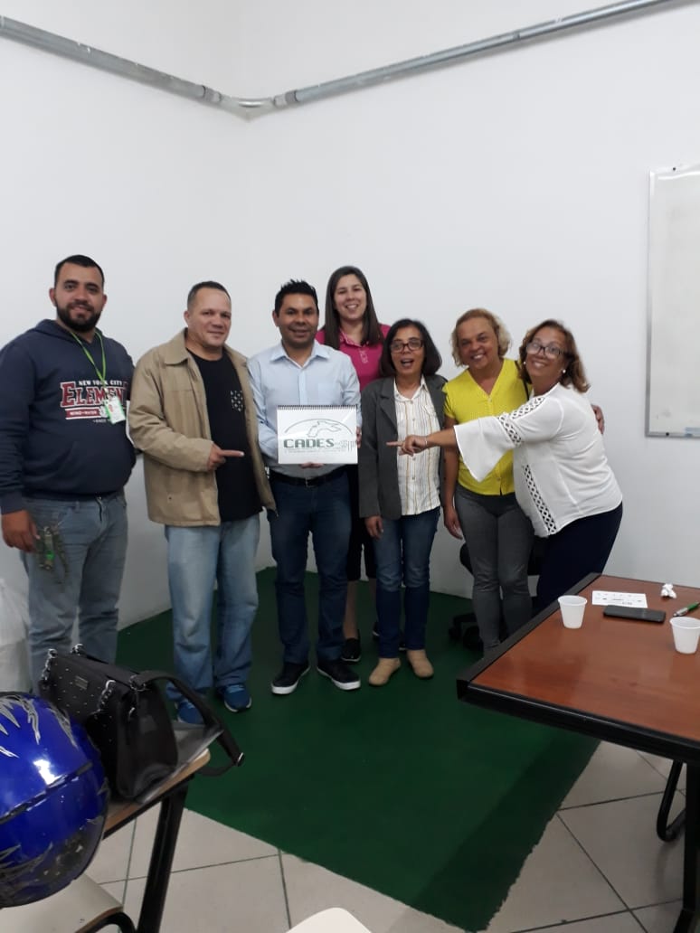 A foto mostra um grupo de sete pessoas adultas em um ambiente interno, posando para a câmera. Eles estão em pé, lado a lado, com um sorriso no rosto, transmitindo uma atmosfera de celebração ou reconhecimento.  Ao centro do grupo, um homem veste uma camisa azul clara e calças jeans escuras. Ele segura uma placa retangular branca com letras pretas que parecem formar uma palavra ou sigla ("CHOUS" é parcialmente visível). As pessoas ao seu redor parecem direcionar o olhar e, em alguns casos, apontar para a placa, indicando que ela é o foco da fotografia.  À esquerda do homem com a placa, há dois homens. O mais à esquerda veste uma blusa de moletom azul escura e calças jeans claras. O homem ao seu lado veste uma jaqueta bege sobre uma camiseta preta e calças jeans azuis claras.  À direita do homem com a placa, há quatro mulheres. A primeira veste uma blusa rosa e calças jeans escuras. A segunda veste um blazer cinza sobre uma blusa estampada e calças jeans azuis. A terceira veste uma camiseta amarela e calças cinzas. A mulher mais à direita veste uma blusa branca de mangas compridas e calças azuis escuras. Ela está com o braço estendido em direção à placa, com um sorriso largo e óculos.  O ambiente ao fundo é uma parede branca com uma faixa verde na parte inferior. À direita, é possível ver parte de um quadro branco. Na frente do grupo, à esquerda, há uma mesa com uma bolsa preta e um capacete azul. À direita, outra mesa com alguns copos brancos e papéis. A iluminação parece artificial, vinda do teto.  A composição da foto sugere um momento de entrega, premiação ou reconhecimento, onde a placa tem um significado especial para o grupo. As expressões faciais e a linguagem corporal das pessoas indicam alegria e união.