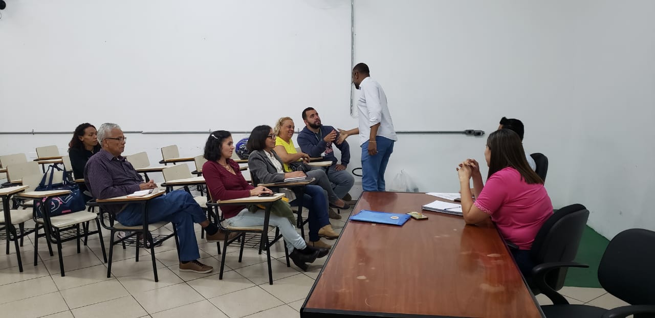 A imagem mostra uma sala de aula com um grupo de sete pessoas sentadas em cadeiras com braços, voltadas para a direita da imagem, onde um homem está em pé, parecendo conduzir alguma atividade ou instrução. As pessoas sentadas parecem estar prestando atenção.  O homem em pé veste uma camisa branca de mangas compridas e calças jeans azuis. Ele está de perfil, voltado para as pessoas sentadas, e parece estar gesticulando com as mãos enquanto fala ou demonstra algo.  As pessoas sentadas são diversas em aparência. Da esquerda para a direita, observa-se:  Uma mulher com cabelos escuros e longos, vestindo uma blusa clara e calças jeans escuras. Um homem com cabelos grisalhos e barba rala, vestindo uma camisa de manga comprida vinho e calças jeans azuis. Uma mulher com cabelos escuros e curtos, vestindo uma blusa vinho e calças claras. Uma mulher com cabelos claros, vestindo uma jaqueta cinza sobre uma blusa amarela e calças jeans. Um homem com barba e cabelos escuros, vestindo uma camiseta azul clara e calças jeans. Uma pessoa com cabelos escuros e óculos, vestindo uma camiseta rosa. A sua posição dificulta a identificação do gênero com certeza. Uma pessoa com cabelos escuros, vestindo uma camiseta rosa, sentada de costas para a câmera em uma mesa separada, parecendo estar envolvida na atividade. A sala de aula tem paredes brancas com uma faixa horizontal mais escura na parte inferior. O chão é revestido por ladrilhos brancos. As cadeiras dos alunos são brancas com estrutura metálica preta. À direita, há uma mesa de cor marrom com alguns papéis e uma pasta azul sobre ela. A iluminação parece ser artificial, vinda do teto.  A atmosfera da imagem sugere um ambiente de aprendizado ou treinamento, com um instrutor conduzindo uma sessão para um grupo de participantes.