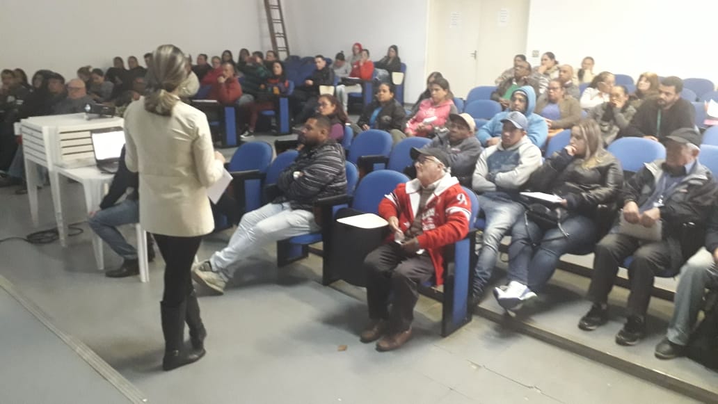 A foto mostra uma sala de aula ou auditório com um grande grupo de pessoas sentadas em cadeiras azuis, assistindo a uma apresentação. Uma mulher está em pé na frente, de costas para a câmera, parecendo ser a apresentadora.  A apresentadora veste um casaco claro, calças e botas escuras. Ela parece estar falando ou gesticulando, possivelmente utilizando um notebook branco que está aberto sobre uma mesa branca à sua esquerda.  A plateia é composta por diversas pessoas, preenchendo quase todas as cadeiras disponíveis. As pessoas estão voltadas para a apresentadora, e algumas parecem estar olhando para ela atentamente. É possível observar diferentes vestimentas e cortes de cabelo entre os participantes.  A sala tem paredes brancas e o chão parece ser de concreto ou algum material similar. Ao fundo, à esquerda, há uma escada metálica encostada na parede. A iluminação do ambiente parece ser artificial, vinda do teto.  A atmosfera da imagem sugere um evento educacional, como uma palestra, um curso ou um treinamento, com uma apresentadora compartilhando informações com uma audiência considerável. A sala cheia indica um bom comparecimento ou um tema de interesse para muitas pessoas.