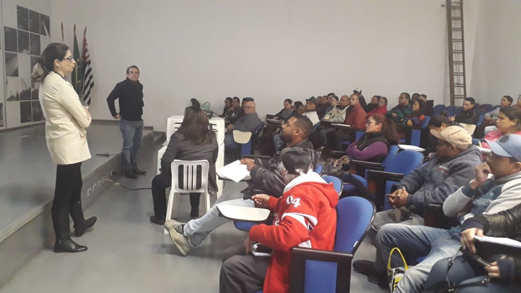 A foto mostra uma sala de aula ou auditório com um grande grupo de pessoas sentadas em cadeiras azuis, assistindo a uma apresentação. Uma mulher está em pé na frente, de costas para a câmera, parecendo ser a apresentadora.  A apresentadora veste um casaco claro, calças e botas escuras. Ela parece estar falando ou gesticulando, possivelmente utilizando um notebook branco que está aberto sobre uma mesa branca à sua esquerda.  A plateia é composta por diversas pessoas, preenchendo quase todas as cadeiras disponíveis. As pessoas estão voltadas para a apresentadora, e algumas parecem estar olhando para ela atentamente. É possível observar diferentes vestimentas e cortes de cabelo entre os participantes.  A sala tem paredes brancas e o chão parece ser de concreto ou algum material similar. Ao fundo, à esquerda, há uma escada metálica encostada na parede. A iluminação do ambiente parece ser artificial, vinda do teto.  A atmosfera da imagem sugere um evento educacional, como uma palestra, um curso ou um treinamento, com uma apresentadora compartilhando informações com uma audiência considerável. A sala cheia indica um bom comparecimento ou um tema de interesse para muitas pessoas.