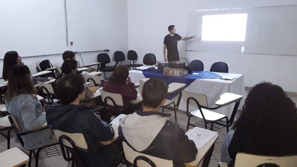 A foto mostra uma sala de aula com um grupo de pessoas sentadas em carteiras individuais, voltadas para a frente, onde um homem está em pé ao lado de uma tela branca, parecendo ser o instrutor ou apresentador.  O instrutor está à direita da imagem, próximo à tela. Ele veste uma camiseta escura e calças compridas escuras. Ele está de perfil, com o braço direito estendido em direção à tela, como se estivesse apontando ou explicando algo.  Os alunos estão sentados em carteiras com acento preto e apoio para escrita branco. Eles estão virados para a frente, observando o instrutor e a tela. É possível identificar pelo menos sete alunos, com diferentes cortes de cabelo e vestimentas. Alguns parecem estar tomando notas.  Na frente da sala, há uma mesa coberta por uma toalha branca e um tecido azul por cima. Há alguns objetos sobre a mesa, mas não são claramente distinguíveis. Atrás da mesa, há algumas cadeiras vazias.  A sala de aula tem paredes brancas e o chão parece ser claro. A iluminação é artificial, vinda do teto. A tela branca na frente sugere o uso de algum tipo de projeção ou é um quadro branco para anotações.  A cena retrata um ambiente de aprendizado, com um instrutor conduzindo uma aula ou apresentação para um grupo de alunos. A disposição da sala e a atenção dos alunos indicam uma interação formal de ensino