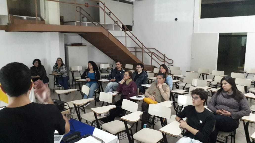 A foto mostra uma sala de aula ou auditório com um grupo de pessoas sentadas em cadeiras com pequenas mesas acopladas, voltadas para a esquerda da imagem, onde um indivíduo parece estar fazendo uma apresentação.  O apresentador está em primeiro plano, de costas para a câmera, vestindo uma camiseta preta e com a parte de trás da cabeça visível, mostrando cabelos escuros. Seus braços estão levemente levantados e as mãos parecem estar gesticulando enquanto fala.  O público é composto por diversas pessoas, sentadas em várias fileiras de cadeiras brancas com estrutura preta e mesas laterais brancas. As pessoas estão olhando na direção do apresentador. É possível observar diferentes vestimentas, cortes de cabelo e poses entre os participantes.  Ao fundo, a sala tem paredes brancas e uma estrutura de mezanino ou segundo andar com uma escada de madeira com corrimão metálico visível no canto superior direito. Há também uma porta escura no fundo, à direita. A iluminação do ambiente parece ser artificial, vinda do teto.  A atmosfera da imagem sugere um ambiente de aprendizado, como uma palestra, um seminário ou uma aula, com um apresentador compartilhando informações com um público atento. A disposição das cadeiras e a postura das pessoas indicam uma interação formal de comunicação.