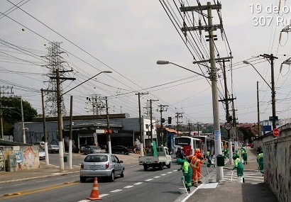 Funcionários de uniforme verde e outros de uniforme laranja trabalham na pintura de guias em uma avenida. Usada tinta branca.
