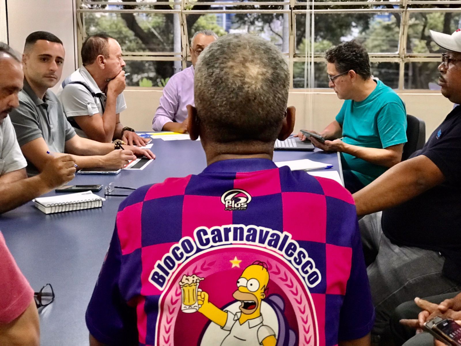 Reunião de um grupo de pessoas ao redor de uma mesa e um deles, em destaque com uma camiseta de estampa carnavalesca