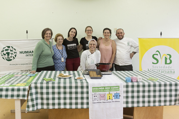 Sete pessoas atrás de uma mesa, sendo dois cozinheiros, dois membros da SMADS e três pessoas da equipe da SVB. Em cima da mesa estão alimentos e um pequeno fogão com uma panela.