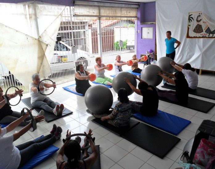 Mulheres sentadas em colchonetes se alongando com bambolês e bolas.